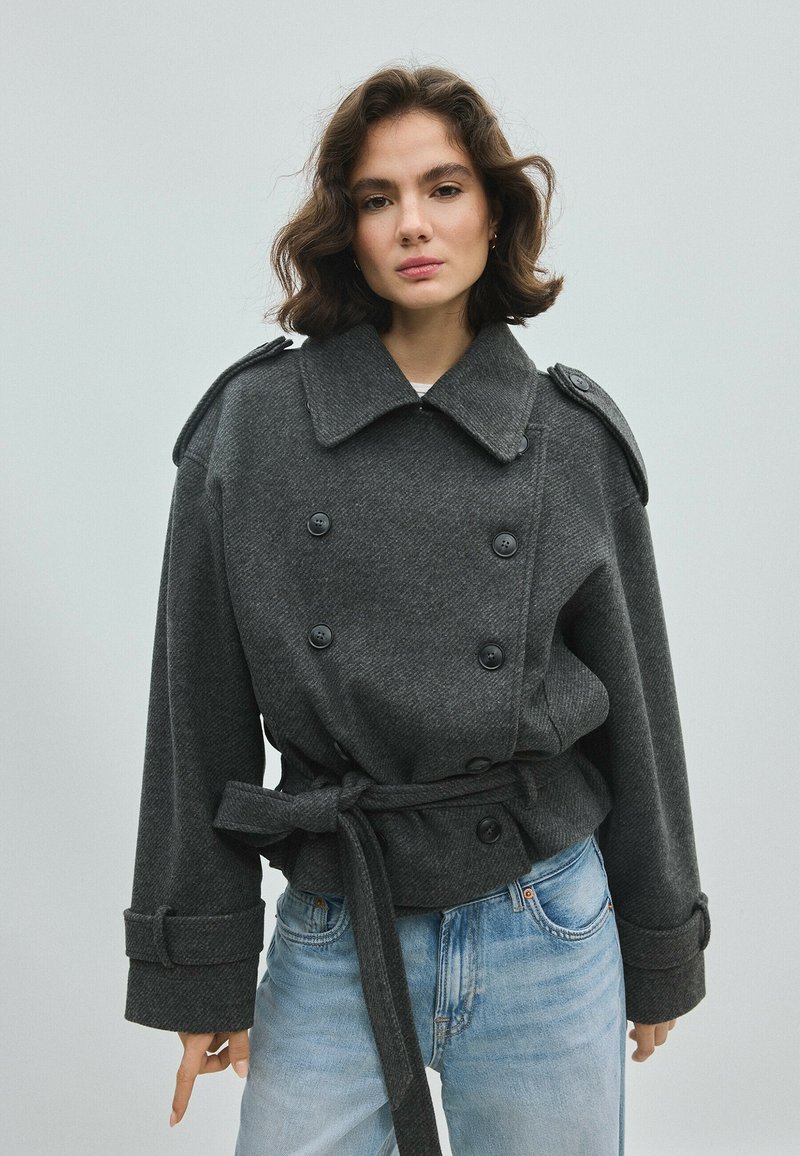 Femme aux cheveux courts et ondulés porte une veste à double boutonnage ceinturée gris foncé et un jean bleu clair sur un fond clair uni.