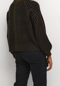 Maglione marrone con strisce diagonali blu, scollo a coste e spalle basse; abbinato a jeans neri aderenti. Tessuto morbido e vestibilità rilassata.