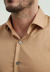 Chemise boutonnée beige clair avec une texture douce, dotée d'un col classique et de deux boutons gris à l'avant.