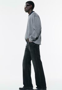 Maglione grigio a costine con colletto nero, abbinato a jeans a gamba larga in denim scuro e scarpe nere. La persona indossa occhiali da sole.
