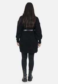 Cardigan en tricot noir surdimensionné avec une texture côtelée, ceinturé à la taille avec une sangle en cuir noir, associé à des leggings noirs et des bottes.