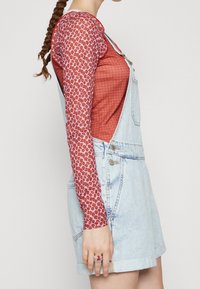 Helleblaue Jeanslatzhose kombiniert mit einem langärmeligen rot gemusterten Oberteil. Der Latzhose hat verstellbare Träger und Seitentaschen.