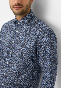 Camicia abbottonata di colore blu scuro con un motivo geometrico blu chiaro e marrone. Presenta un colletto aperto e bottoni bianchi.