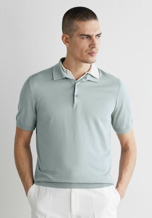 Mann in hellgrauem kurzärmligem Poloshirt mit weißem Kragen und hellen Hosen, stehend vor einer weißen Wand, Hände in den Taschen.