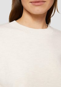 Hellbeige Pullover mit rundem Halsausschnitt, hergestellt aus weichem, strukturierter Stoff und mit geripptem Kragen. Nahaufnahme zeigt die Nähte und subtile Stoffmuster.