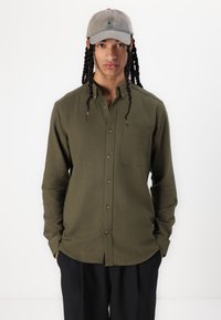 Camisa verde oliva de botones con un bolsillo en el pecho, tela texturizada y dobladillo curvo, llevada con una gorra gris y pantalones negros.