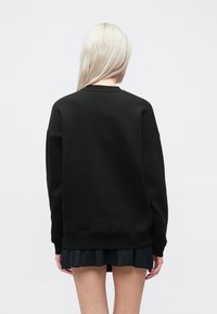 Melna lielgabarīta kapuces džemperis ar pazeminātiem pleciem, kokvilnas materiāls un ribota apakšmala, kombinēts ar plīsu melnu svārku.