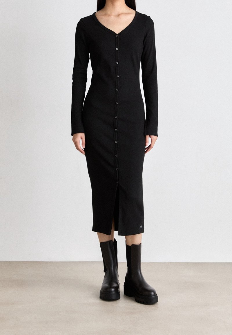 Femme portant une robe midi noire cintrée à manches longues avec boutons, associée à des bottes noires épaisses mi-mollet, debout sur un sol uni.
