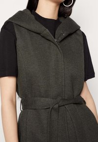 Ärmelloses Oberbekleidungsstück aus dunkelgrünem Stoff mit strukturierter Oberfläche, ausgestattet mit Kapuze, Reißverschluss und tailliertem Gürtel für eine verstellbare Passform.