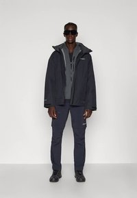 Μαύρο αδιάβροχο μπουφάν με κουκούλα, το οποίο είναι φορεμένο πάνω από έναν γκρίζο τοπ, συνδυασμένο με σκούρες cargo παντελόνες και μαύρα παπούτσια, με ανακλαστικές λεπτομέρειες.