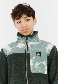 Fleecejack met groene textuurstof, een gladde lichtgroene bovenkant en een witte spetterpatroon. Voorzien van een ritszak en hoge kraag.