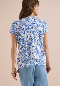 Mujer con camiseta blanca de manga corta con estampado azul de paisley, combinada con shorts vaqueros azul claro, de pie frente a un fondo beige liso.
