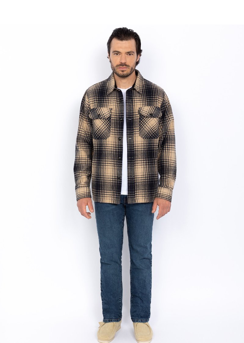 Camicia flanella a quadri beige e nera con due tasche sul petto, indossata sopra una t-shirt bianca. Abbinata a jeans blu e scarpe beige.