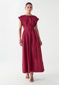 Burgundská maxi šaty s krátkymi rukávmi, sťahovaným pásom a viazaním. Má jemnú textúru a skladanie, nosené s béžovými sandálmi.