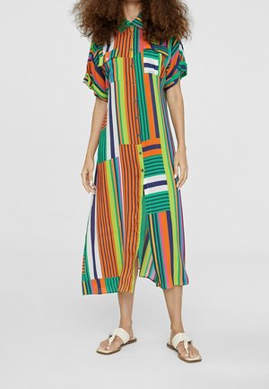 Kvinde iført en farverig, stribet midikjole med knapper, korte ærmer og forlommer, kombineret med off-white flade sandaler.