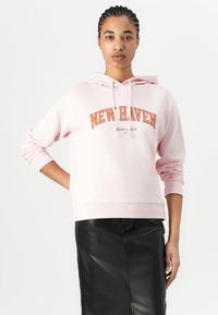 Felpa con cappuccio rosa chiaro con la scritta "NEW HAVEN" in lettere arancioni. Presenta una vestibilità comoda e maniche lunghe. Abbinata a una gonna in pelle nera.