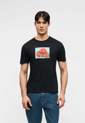 Άνδρας που φοράει μαύρο T-shirt με γραφικό από μια κόκκινη καλυμμένη σκούτερ και τη λέξη "HUGO" στο μπροστινό μέρος, συνδυασμένο με μπλε τζιν.