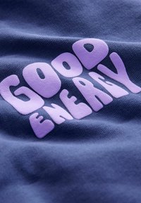 Tessuto navy con una superficie texturizzata caratterizzato da grandi lettere rotonde di colore viola chiaro che scrivono "GOOD ENERGY" in modo prominente sul davanti.