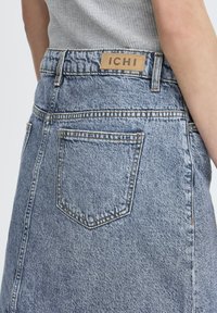 Jupe en denim avec taille haute, lavage bleu, poches arrière et une étiquette beige marquée "ICHI" sur la ceinture. Détails de tissu texturé visibles.