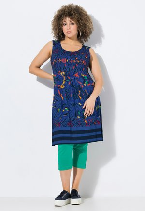 Ärmelloses blaues Kleid mit einem bunten abstrakten Muster und horizontalen Streifen am Saum, kombiniert mit grünen Caprihosen und schwarzen Turnschuhen.