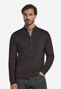 Mörkbrun zip-up cardigan i mjukt tyg, med ståkrage, ribbade ärmslut och en slim fit-design. Kombinerad med en beige skjorta.