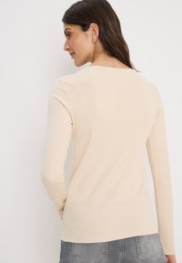 Haut beige à manches longues avec un col rond. Tissu lisse et extensible, avec une silhouette ajustée. La vue arrière montre un design uni et des coutures nettes.