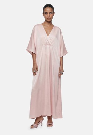 Nainen pukeutuneena väljään mahdollisesti vaaleanpunaiseen satiiniseen maximekkoon, jossa on leveät hihat ja V-pääntie, yhdistettynä kimaltaviin pinkkeihin avonaisiin korkokenkiin ja hopeisiin sormuksiin.