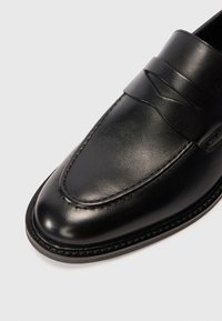 Melnas ādas loafer kurpes ar noapaļotu purngalu, ar izteiksmīgu šuvju detaļu un dekoratīvu siksniņu pāri augšdaļai, uz balta fona.