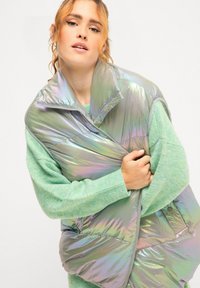 Iridescent opblaasbare vest met een hoge kraag, met een glanzende textuur en ritssluiting, gelaagd over een lichtgroene gebreide trui.