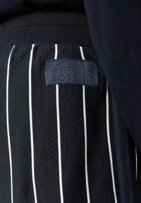 Zwarte stof met verticale witte krijtstrepen, voorzien van een gestikt patch met een basketbalafbeelding, de tekst "BOSS" en het jaar "1977".