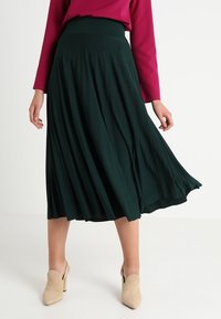 Gonna midi plissettata verde realizzata in tessuto leggero con vita alta. Abbinata a una maglietta magenta a maniche lunghe e scarpe beige.