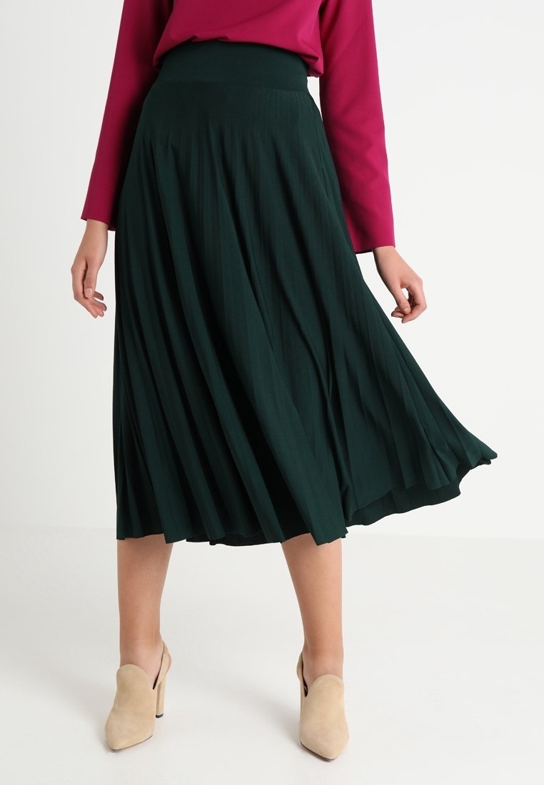 Gonna midi plissettata verde realizzata in tessuto leggero con vita alta. Abbinata a una maglietta magenta a maniche lunghe e scarpe beige.