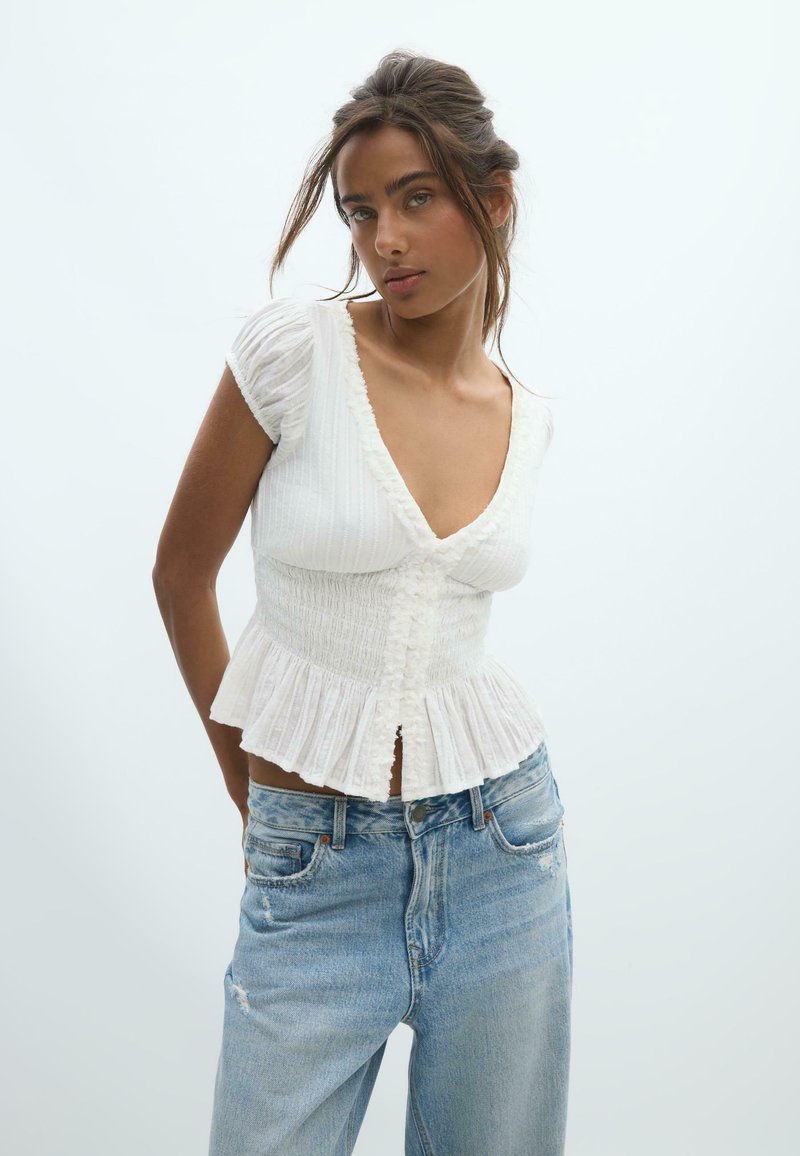 Woman with brown hair in a white short-sleeve ruffled blouse and light blue high-waisted jeans, standing against a plain light background.
