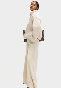Kurtka polarowa na zamek błyskawiczny w jasnobeżowym kolorze, z wysokim kołnierzem i oversize'owym krojem. W połączeniu z szerokimi, kremowymi spodniami i średniej wielkości torebką w odcieniu średniego brązu.