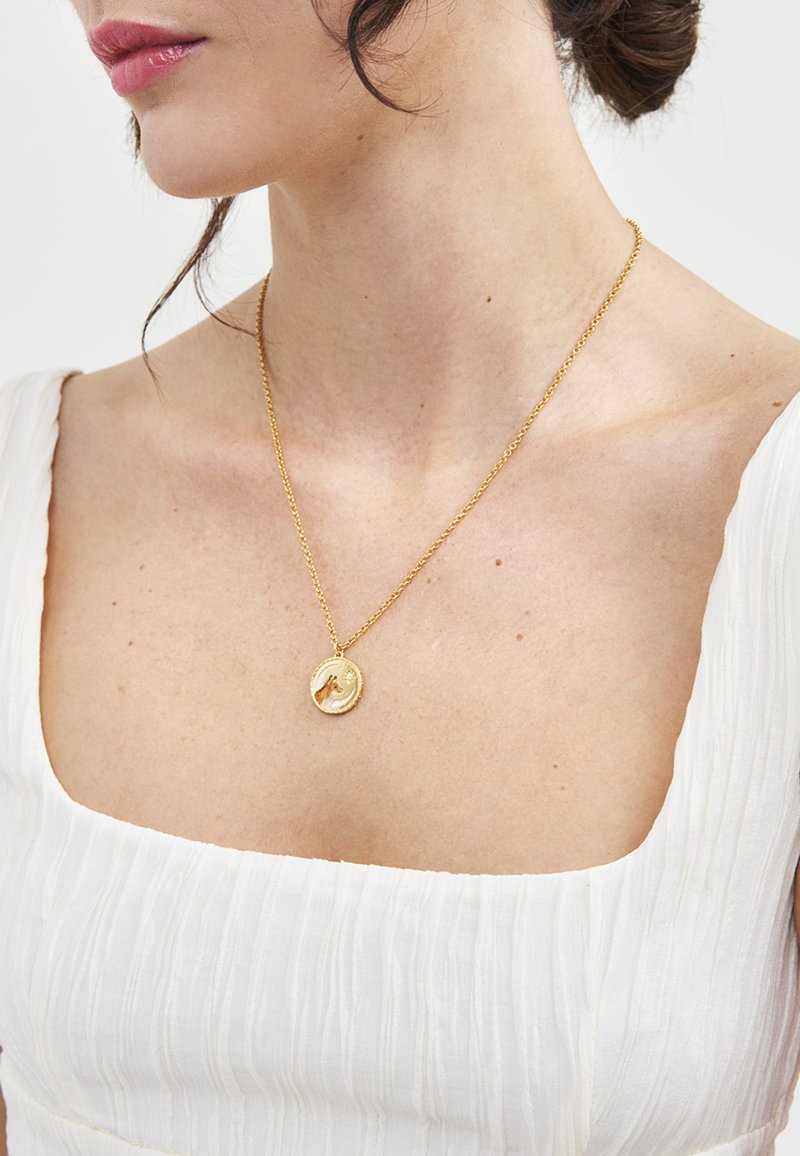 Close-up of a woman wearing a white square-neck top and a gold pendant necklace with a hand design.