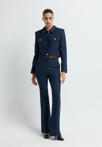 Námořnicky modrá krátká bunda se stříbrnými knoflíky, dvě přední kapsy; v kombinaci s odpovídajícími vysokými zvonovými kalhotami. Hladká textura látky.