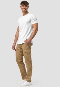 T-shirt blanc à col rond, pantalon cargo beige avec poches latérales, et baskets blanches, présentés sur une figure masculine debout devant un fond gris.