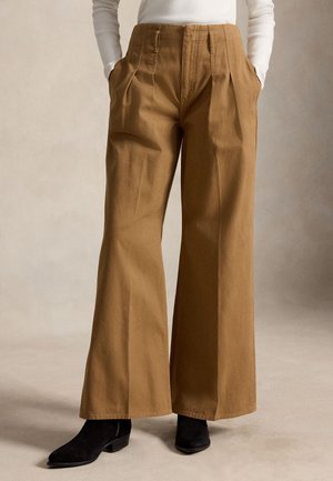 Braune Hose mit hohem Bund und weitem Bein mit vorderen Falten, kombiniert mit schwarzen Stiefeletten und einem in die Hose gesteckten weißen Top. Hände in den Taschen.