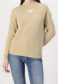 Beige geribbelde trui met een hoge halslijn, voorzien van een logopatch aan de voorkant, gecombineerd met blauwe spijkerbroek.