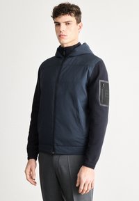 Tumši zila kapuce jaka ar rāvējslēdzēja aizdari, kontrastējošām piedurknēm un rāvējslēdzēja kabatu uz kreisās rokas. Gluda tekstūra un mūsdienīgs piegriezums.