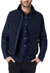 Marineblauer, gestepptes Weste mit hohem Kragen, ausgestattet mit einem Reißverschluss und strukturiertem Muster, getragen über einem dunklen Denim-Hemd, das Taschen und Knöpfe hervorhebt.