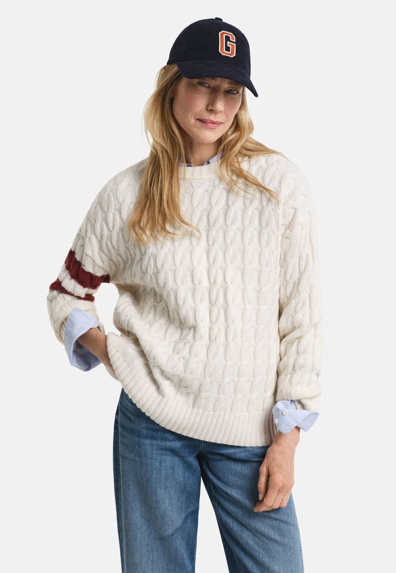 Kabelstrick-Creme-Pullover mit rotem Streifenakzent und geripptem Saum, kombiniert mit einem hellblauen Hemd mit Kragen und einer marineblauen Baseballmütze mit dem Buchstaben "G."