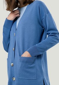 Modrý pletený cardigan s výstřihem do V, předními knoflíky, žebrovanými manžetami a dvěma předními kapsami. Měkká textilie se bočními rozparky pro pohodlné nošení.