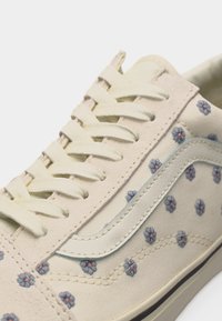 Baskets couleur crème avec lacets blancs, fleurs bleues brodées et bande latérale blanche sur une semelle de couleur claire.