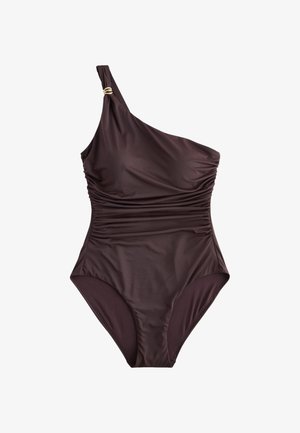Kvinde iført en svart enkelt skulder badedragt og gyldne armbånd står ved en stor sten på en sandstrand med grønt i baggrunden.