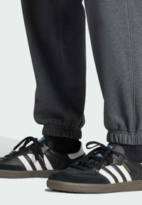 Svarta och grå sportskor med vita ränder, en texturerad överdel av läder och mocka, samt en gummisula. Svarta elastiska byxor med muddar är delvis synliga.