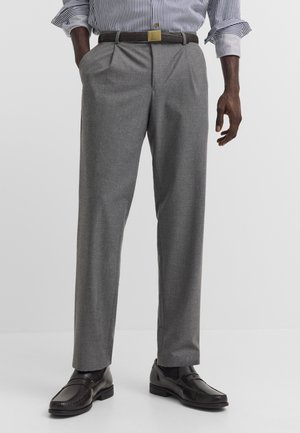 Mann trägt graue Anzughose, schwarzen Gürtel mit goldener Schnalle, schwarze Leder-Loafer und ein in die Hose gestecktes blau-weiß gestreiftes Hemd mit hochgekrempelten Ärmeln.