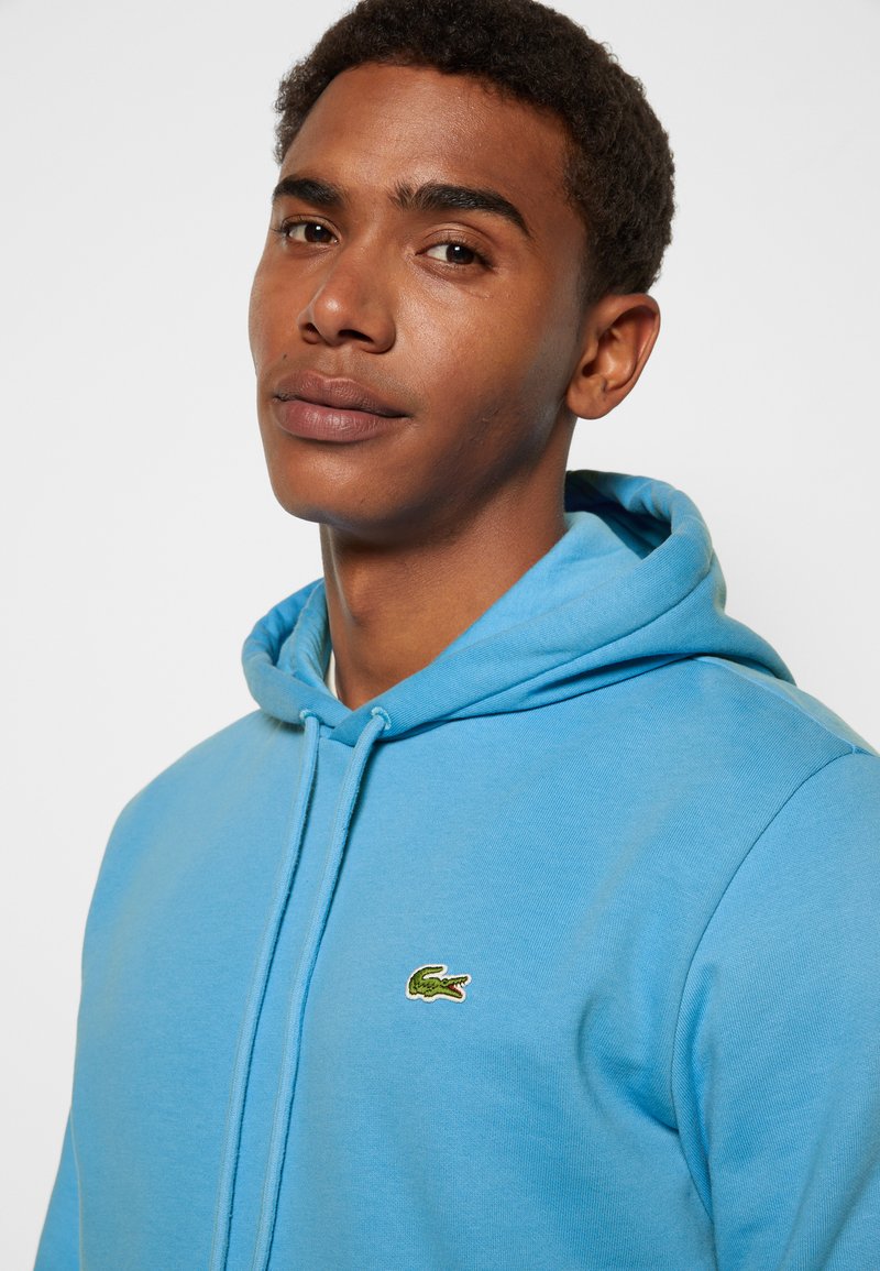 Sweatshirt bleu clair avec capuche, arborant un petit logo de crocodile vert sur la poitrine. Tissu doux, design décontracté et coupe standard.