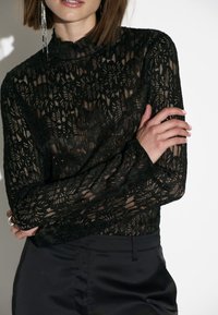 Schwarze Spitzenbluse mit hohem Kragen und langen Ärmeln, verziert mit floralen Mustern und einer strukturierten Oberfläche, kombiniert mit eleganten schwarzen Hosen.