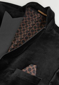 Zwarte fluwelen blazer met een grijze revers. Aan de binnenkant heeft het een geometrisch patroon in donkere en koperen tinten. De pochet past bij het interieurontwerp.